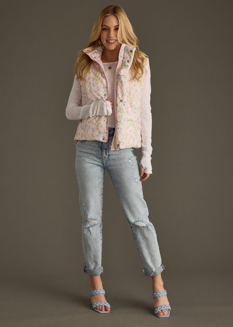 May include: A pink floral print puffer vest worn over a white long-sleeved shirt and light blue jeans. The person is wearing light blue sandals.