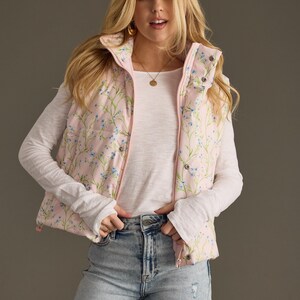 May include: A pink puffer vest with a floral print featuring small blue flowers. The vest has a zip closure and a stand-up collar.