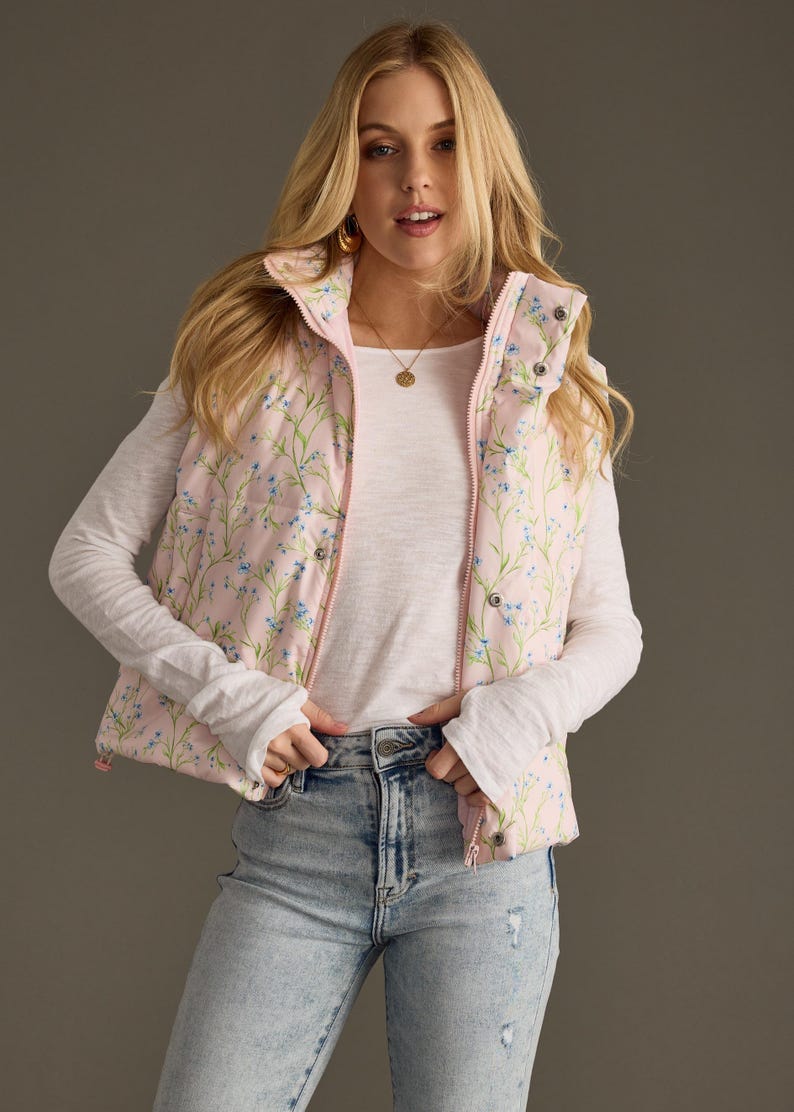 May include: A pink puffer vest with a floral print featuring small blue flowers. The vest has a zip closure and a stand-up collar.