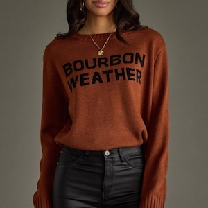 May include: A rust-colored sweater with the text "BOURBON WEATHER" in black block letters. The sweater is paired with black pants. The model is wearing a gold necklace and rings.