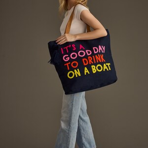 May include: Navy blue tote bag with the embroidered phrase "IT'S A GOOD DAY TO DRINK ON A BOAT" in pink, orange, and yellow. The bag has a brown leather handle and a navy tassel. A casual accessory for a day out.