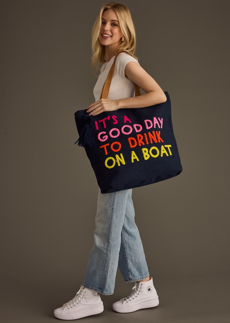 May include: Navy blue tote bag with the embroidered phrase "IT'S A GOOD DAY TO DRINK ON A BOAT" in pink, orange, and yellow. The bag has a brown leather handle and a navy tassel. A casual accessory for a day out.