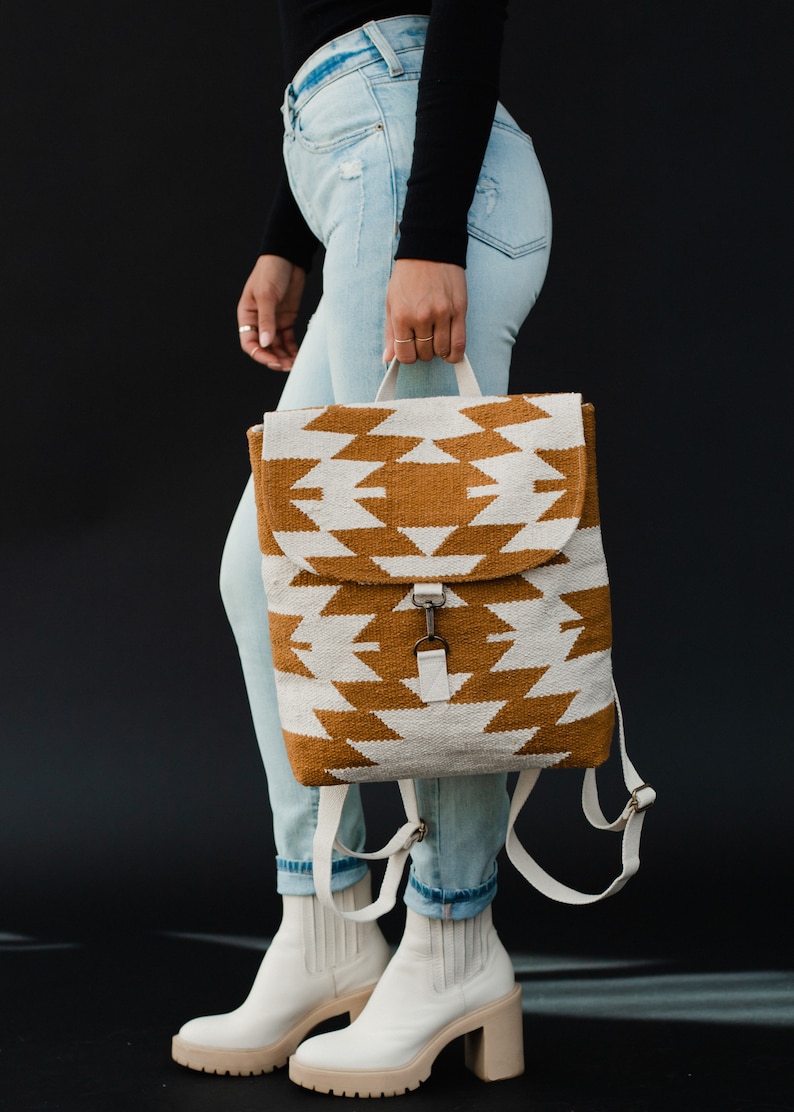 May include: A white and brown patterned backpack with a white canvas strap. The backpack is being held by a person wearing blue jeans and white ankle boots.