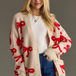 May include: A beige cardigan sweater with red bow accents. The open-front sweater has two pockets and is buttoned. The model is wearing a white top and blue jeans. The sweater is a cozy and stylish addition to any wardrobe.