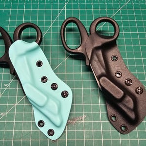 May include: Two tactical shears with protective sheaths. One sheath is a light teal color, and the other is black. Both sheaths have multiple black rivets. The shears have large finger holes and a curved blade.