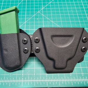 May include: A black leather holster with a green magazine holder attached. The holster has multiple screws and is designed to be worn on a belt.