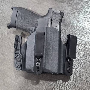 May include: A black handgun in a black holster. The handgun has a textured grip and a ridged slide. The holster has a belt clip and is secured with screws. The image is taken from an overhead perspective.