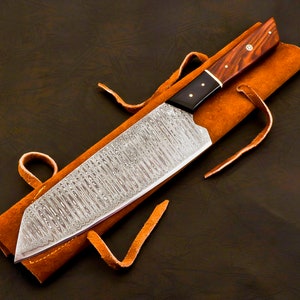 May include: A Damascus steel chef's knife with a wooden handle and a leather sheath. The blade has a unique pattern, and the handle is a dark wood with brass rivets. The sheath is a light brown color with leather ties.