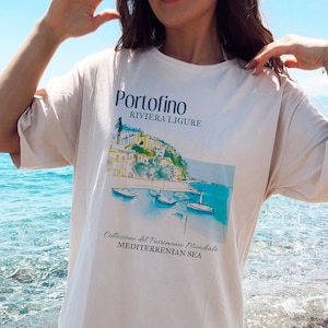May include: A white t-shirt with a graphic print of a seaside town with the text "Portofino Riviera Ligure" and "Collezione del Patrimonio Mondiale MEDITERRENIAN SEA".