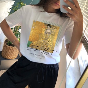 May include: White t-shirt featuring a print of Gustav Klimt's "Adele Bloch-Bauer I" painting. The artwork is in gold, black, and green. The shirt also has the artist's name and the painting's title printed on it.