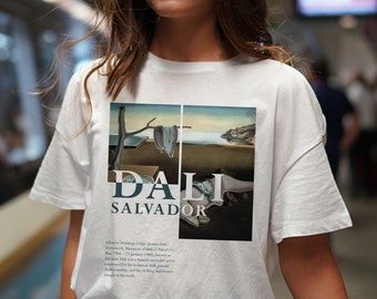 May include: White t-shirt with a graphic print of Salvador Dali's "The Persistence of Memory" painting. The text "DALI SALVADOR" is printed below the image. The text below the image reads: "Salvador Domingo Felipe Jacinto Dali (May 11, 1904 – 23 January 1989), known as Salvador Dali, was a Spanish surrealist artist renowned for his technical skill, and bizarre dreamlike imagery, and the striking and bizarre images in his work."