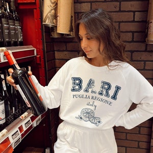 May include: White sweatshirt with a blue and white graphic that says "Bari Puglia Regione Italy" with a lemon design. The sweatshirt is being held by a person in a store.