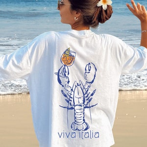May include: White oversized t-shirt with a blue illustration of a lobster holding a glass of orange juice with a slice of lemon. The text "viva italia" is printed below the lobster.