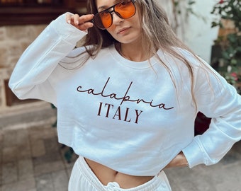 May include: A white cropped sweatshirt with the text "calabria ITALY" printed in maroon on the front. The model is wearing the sweatshirt with a pair of orange-tinted sunglasses.