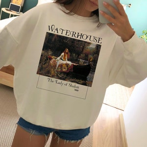 May include: White sweatshirt with a graphic print of a painting by John William Waterhouse titled "The Lady of Shalott" from 1888. The painting depicts a woman in a boat on a river.