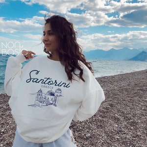 May include: White sweatshirt with a blue graphic of a Santorini, Greece cityscape and the text "Santorini Greece".