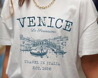 May include: White t-shirt with a blue graphic print of a bridge in Venice, Italy. The text "VENICE La Serenissima TRAVEL IN ITALIA EST. 1928" is printed on the shirt.