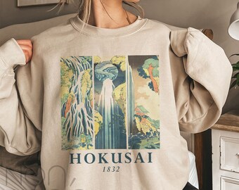 May include: A beige sweatshirt with a vintage Japanese woodblock print of a waterfall. The print is in shades of blue, green, and red. The text "HOKUSAI 1832" is printed below the image.