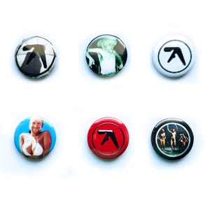 Puede incluir: Seis botones redondos con diferentes imágenes. Los botones son negros, blancos, rojos y azules. Las imágenes incluyen una mujer en bikini, una mujer con un vestido blanco, un logotipo en blanco y negro, un logotipo rojo, un logotipo blanco y una imagen en blanco y negro de tres mujeres.