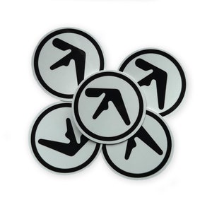 Könnte beinhalten: Eine Ansammlung von fünf runden Aufklebern. Jeder Aufkleber zeigt ein schwarzes, stilisiertes Logo in einem weißen Kreis mit schwarzem Rand. Das Logo ist ein abstraktes Design mit einer einzigartigen Form. Die Aufkleber sind auf weißem Hintergrund angeordnet.