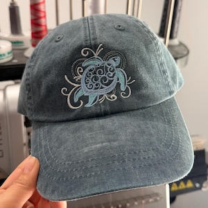 May include: A blue-grey baseball cap featuring an embroidered design of a sea turtle with intricate white and light blue swirling patterns on the front panel. The cap has a curved brim and a washed fabric texture with visible stitching details.