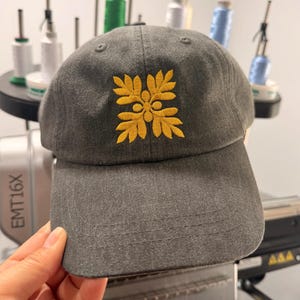May include: A charcoal gray baseball cap featuring a centered, embroidered yellow floral-inspired emblem. The cap has a structured crown, a curved brim, and visible stitching details. The design consists of four symmetrical leaf-like shapes.