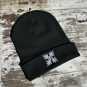 May include: A dark green beanie with a white embroidered snowflake design on the folded brim. The hat is made of a soft, knitted material and is suitable for cold weather. The beanie is displayed on a weathered wooden surface.