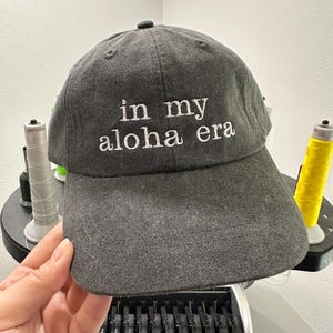 May include: A dark gray baseball cap with white embroidery that reads "in my aloha era".