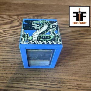 May include: A blue plastic deck box featuring a top panel with a graphic illustration of a green and white snake in a landscape. The front of the box has a clear window displaying a trading card. A logo with the text Ford's Foundry is visible.