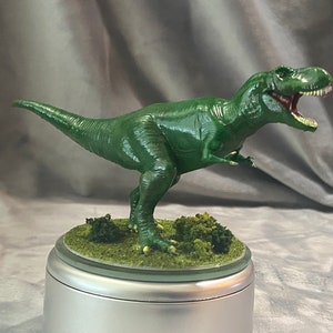 Puede incluir: Figura verde de Tyrannosaurus Rex con la boca abierta, revelando dientes blancos y un interior rojo. El dinosaurio está posado sobre una pequeña base redonda con hierba verde y arbustos. La figura es una representación detallada.