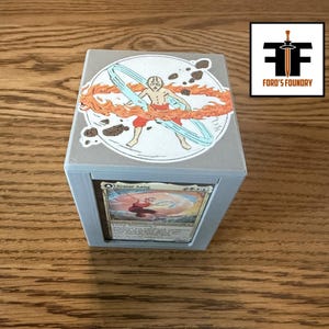 Peut inclure: Une boîte de rangement de cartes cubique grise avec un autocollant circulaire sur le dessus représentant un personnage entouré d'éléments de feu et d'eau. La face avant affiche une carte à collectionner Avatar Aang dans un cadre, avec le logo Ford's Foundry.