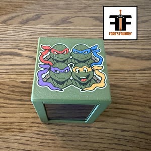 May include: A green cube-shaped box featuring a graphic of four cartoon turtles wearing red, blue, purple, and orange masks on the top surface. The box has a dark rectangular panel on the side and a logo reading Ford's Foundry in the corner.