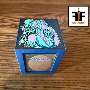 May include: A blue square deck box featuring a top panel with a graphic illustration of a purple-skinned elf with long teal hair. The front panel displays a Magic The Gathering card. A logo in the corner reads Ford's Foundry with a sword icon.