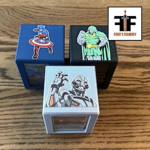 May include: Three square storage boxes featuring comic book character decals. One blue box displays Captain America, one black box features Doctor Doom, and one silver box shows Ultron. Each box has a clear front panel revealing contents inside.