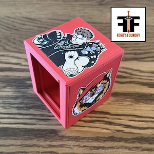May include: A red hollow cube featuring a sticker on the top surface depicting a character with a spiked gauntlet and a sticker on the side surface showing a tiger face with the text THIS IS AN OUT RAGE. The cube has an open front panel.