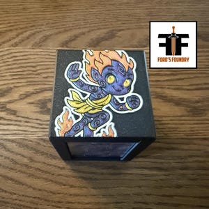 May include: A black cube featuring a graphic illustration of a purple, flame-haired character with yellow eyes and a yellow loincloth. The character is depicted in a dynamic pose surrounded by orange flames. The cube has a textured matte finish.