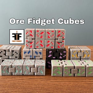 Puede incluir: Un conjunto de ocho cubos fidget, cada uno con un diseño diferente de mineral de Minecraft. Los cubos están hechos de plástico gris y tienen una superficie texturizada. Los diseños incluyen redstone, lapislázuli, hierro, oro, diamante, esmeralda, carbón y obsidiana.