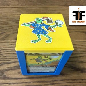 May include: A yellow and blue plastic deck box featuring a sticker of a green frog character wearing a blue tunic and carrying a bindle on its top. The front of the box has a clear window displaying a trading card titled Blobs, the Fool.