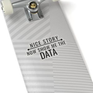May include: A skateboard deck with a gray and white geometric pattern. The deck features the text "NICE STORY NOW SHOW ME THE DATA" in black. The skateboard's trucks and wheels are visible at the top.