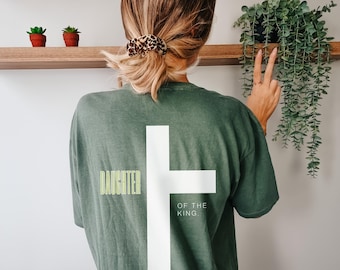 May include: Sage green t-shirt with a large white cross design. The words "DAUGHTER" and "OF THE KING" are printed on the shirt. The word "SAGE" is printed at the bottom of the shirt.