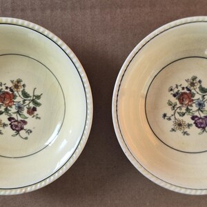 May include: Two round cream-colored ceramic bowls featuring a central floral motif with red, purple, blue, and yellow flowers. Each bowl has a thin dark green circular line accent near the center and a textured, rope-like pattern along the rim.