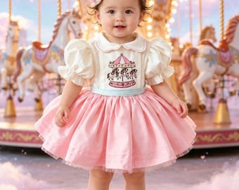 May include: A cream-colored dress with puff sleeves, a Peter Pan collar, and a carousel horse embroidery on the bodice. The dress features a light blue waistband and a full, layered pink skirt. A matching cream hair accessory with a pink bow is worn.