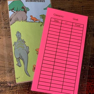 May include: Two planner inserts: one with a dinosaur illustration and the word "DEINONYCHUS", and a bright pink insert with a grid for tracking goals. Both are in clear plastic sleeves.