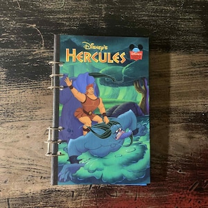 May include: A Disney's Hercules notebook with a spiral binding. The cover features a colourful illustration of Hercules riding a blue monster. The title "Disney's Hercules" is displayed in gold lettering.