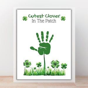May include: White framed art print with the text "Cutest Clover In The Patch". The print features a green handprint with a stem, clover leaves, and grass. The text and handprint are green.