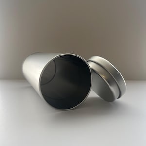 May include: A cylindrical, silver-colored metal container with a matching lid. The container is lying on its side, with the lid slightly ajar, revealing the dark interior. The surface appears smooth and reflective, suggesting a polished finish.