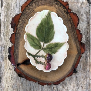 May include: A white ceramic soap dish with a pressed leaf and two red berries. The dish is on a wooden slice.