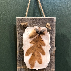 May include: A rustic wooden plaque with a white ceramic embellishment featuring a brown oak leaf and acorns. The plaque has a rope hanger.