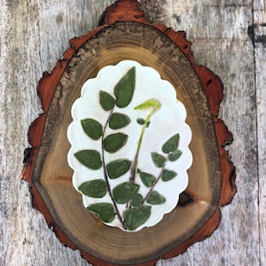 May include: A white ceramic coaster with a green pressed leaf design, sitting on a wooden slice.