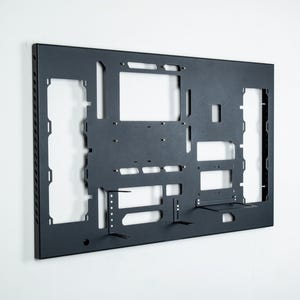 May include: A black metal wall mount with cutouts for mounting computer components. The mount has a textured surface and is designed to be mounted on a wall.